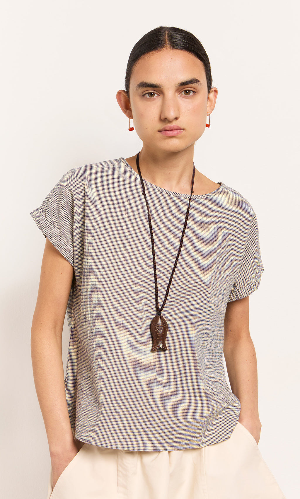 Plümo  Person wearing a short-sleeved top, cream pants, Yumi Fish Necklace, and red earrings stands against a plain wall.