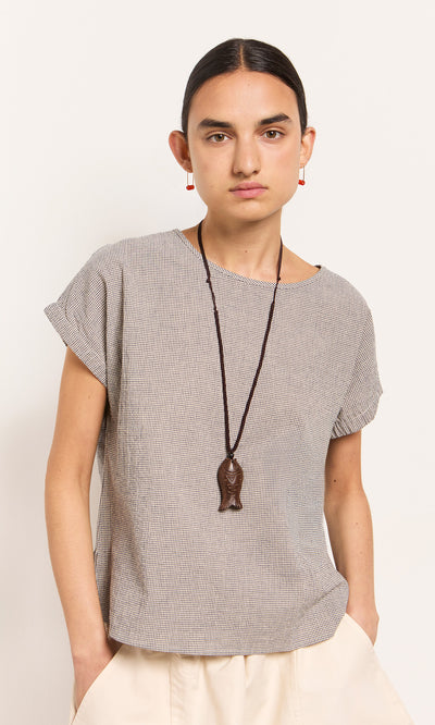 Plümo  Person wearing a short-sleeved top, cream pants, Yumi Fish Necklace, and red earrings stands against a plain wall.