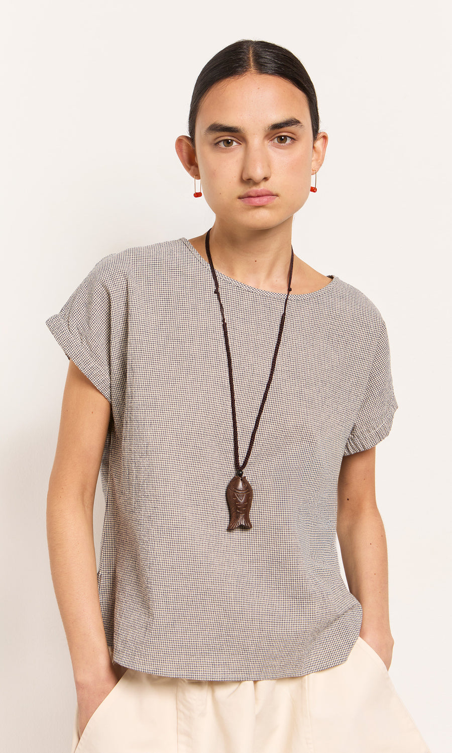 Plümo  Person wearing a short-sleeved top, cream pants, Yumi Fish Necklace, and red earrings stands against a plain wall.