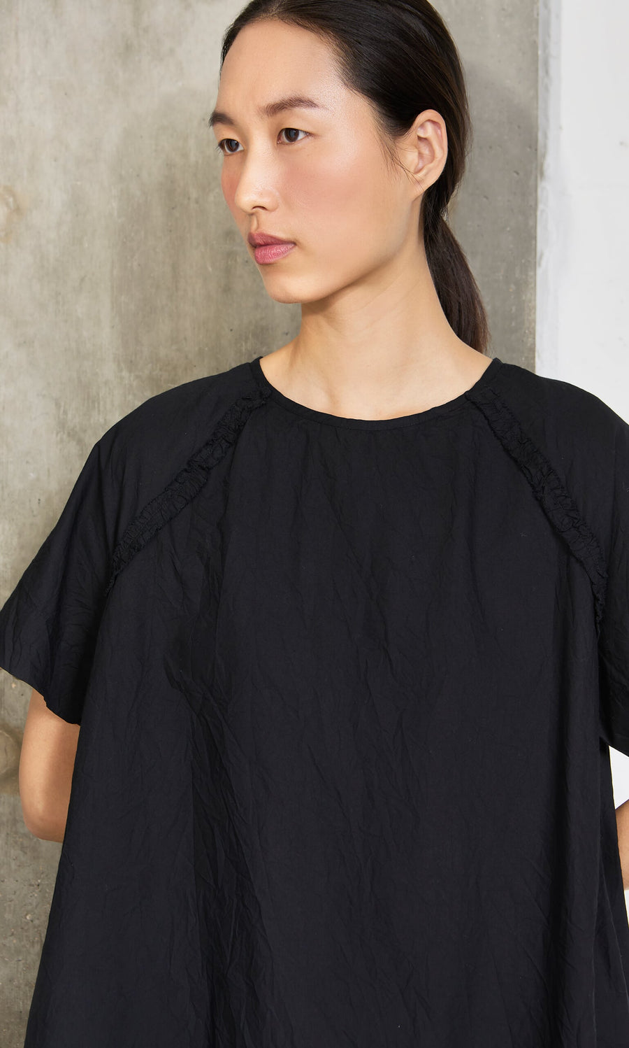 Plümo- Woman with straight dark hair in a ponytail wears the Crinkly Black Blouse, standing by a concrete wall.