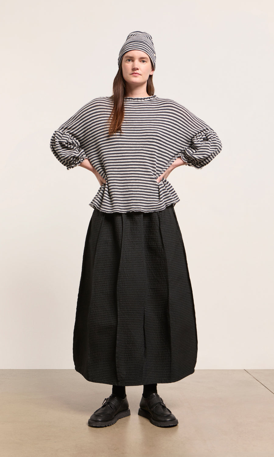 Plümo  Woman wears a striped long-sleeve, Dawn Stripe Beanie, black skirt, socks, and shoes indoors.