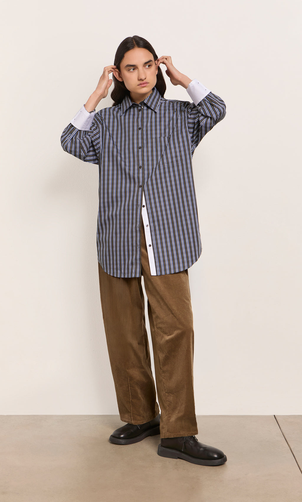 Plümo  Person in a Dubery Shirt and brown pants stands against a plain wall, adjusting their hair.