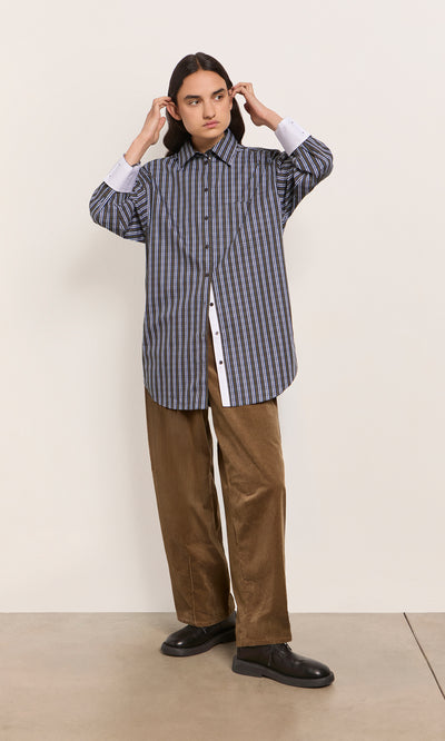 Plümo  Person in a Dubery Shirt and brown pants stands against a plain wall, adjusting their hair.
