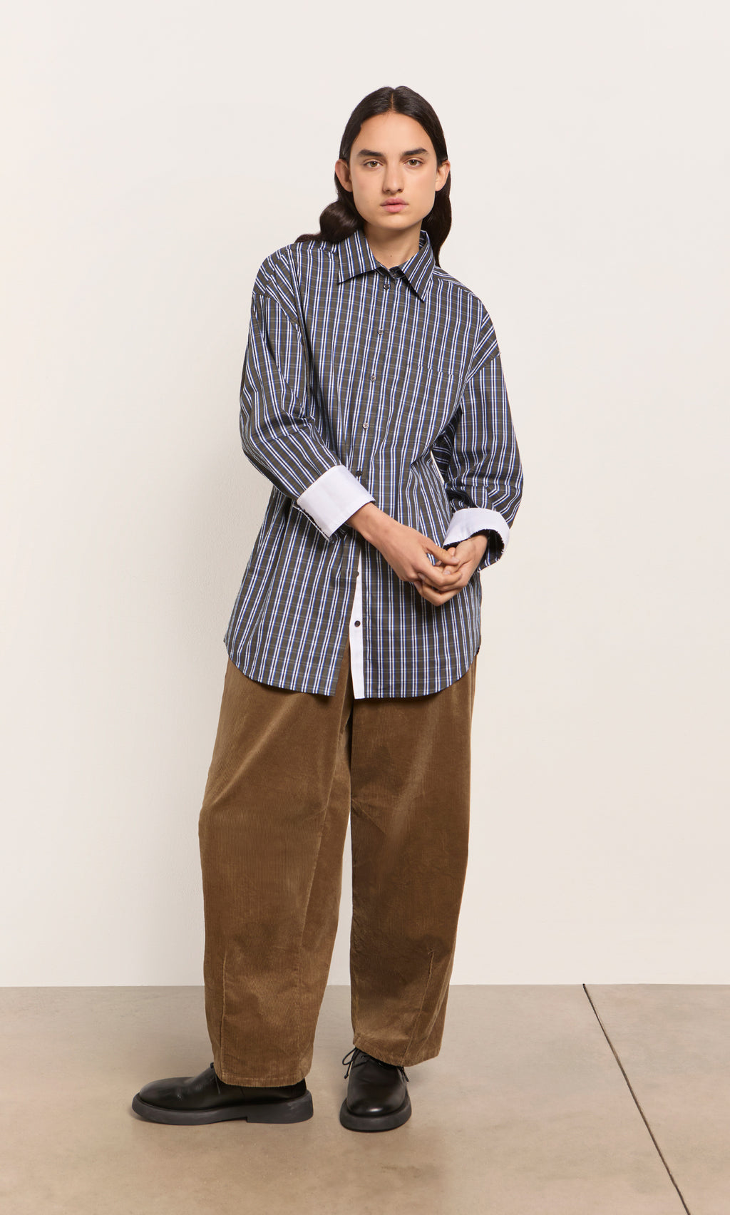 Plümo  Person in a Dubery Shirt, brown corduroy pants, and black shoes stands against a plain wall.