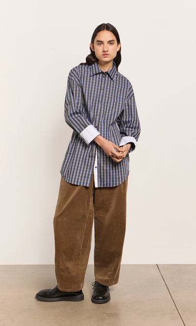 Plümo  Person in a Dubery Shirt, brown corduroy pants, and black shoes stands against a plain wall.