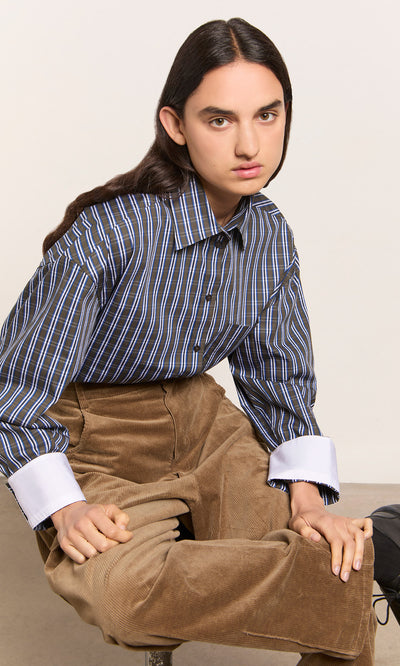 Plümo  Person with long dark hair wears a Dubery Shirt, sitting in tan corduroy pants and looking at the camera.