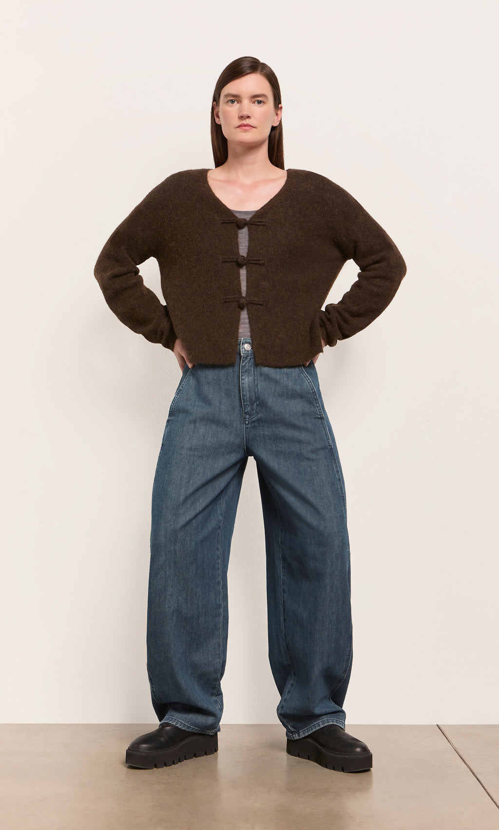 Plümo  Woman with hands on hips wears espresso Duffle Knit Cardigan and wide-leg jeans, channeling Danish style.