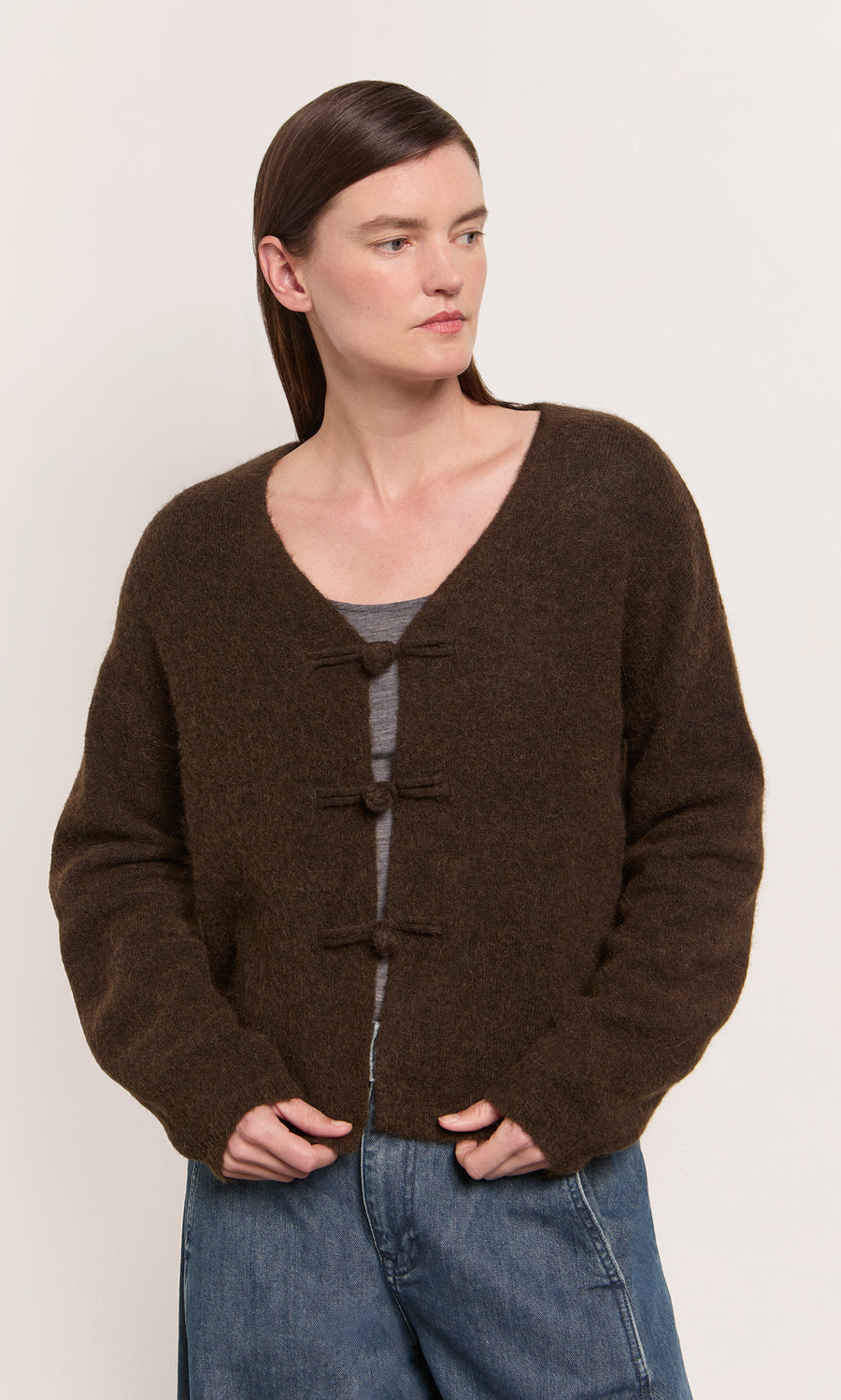 Plümo  Woman in a Duffle Knit Cardigan over a gray top and blue jeans, standing against a light background.