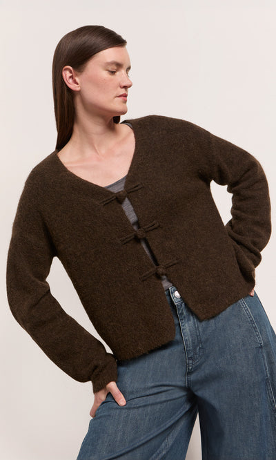 Plümo  Woman in a Duffle Knit Cardigan in espresso chocolate over a gray top and blue jeans, against a light backdrop.