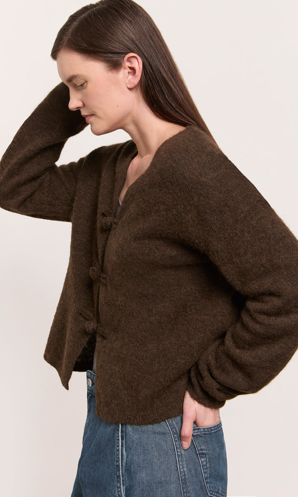Plümo  Woman in a Duffle Knit Cardigan and blue jeans stands sideways, hand on head, against a plain background.