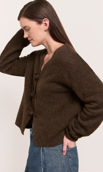 Plümo  Woman in a Duffle Knit Cardigan and blue jeans stands sideways, hand on head, against a plain background.