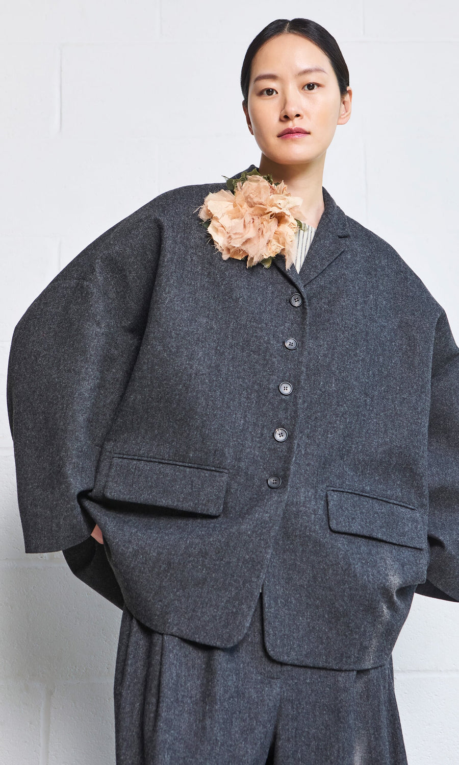 Plümo- A person in an oversized gray suit with a Dusty Rose Corsage stands against a white wall.