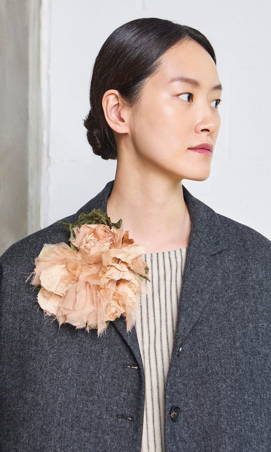 Plümo- Woman with dark hair in a low bun, wearing a gray coat and a Dusty Rose Corsage on her shoulder.
