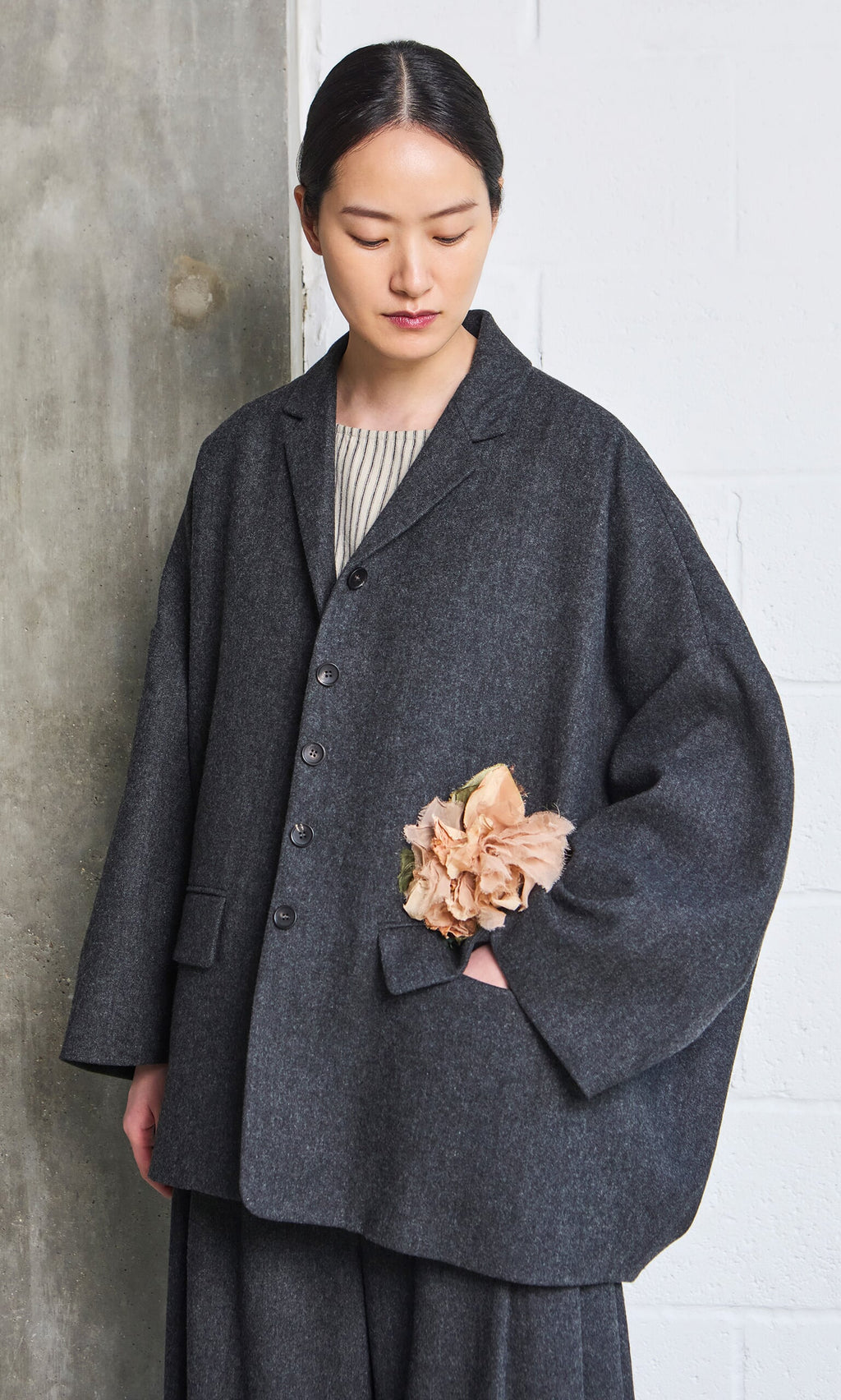 Plümo- A woman in an oversized gray coat stands with a Dusty Rose Corsage in her pocket, looking down.
