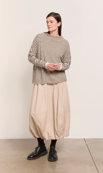 Plümo  Woman in Oana Mock Neck brown striped sweater, beige balloon skirt, black socks and shoes, standing indoors.