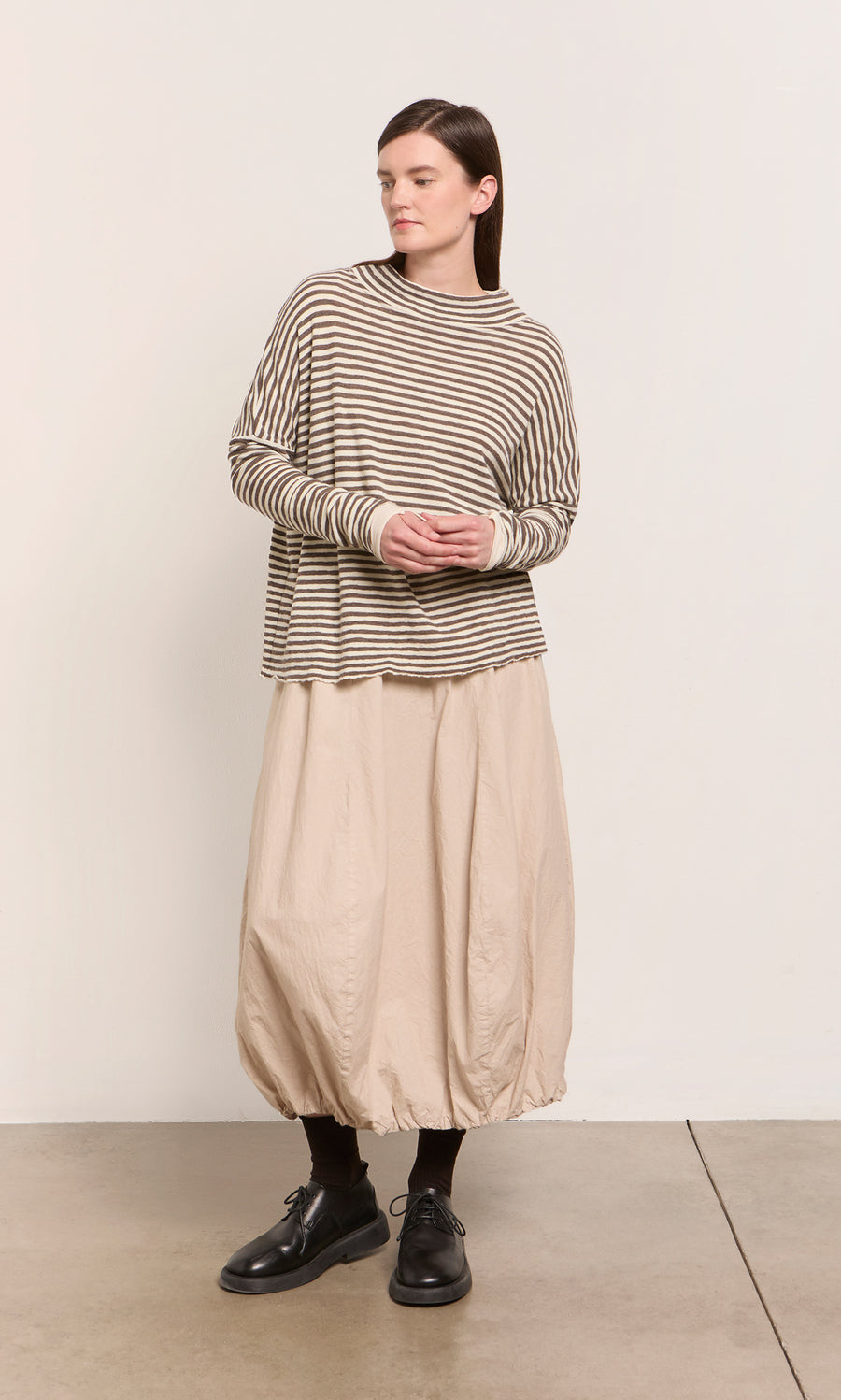 Plümo  Woman in Oana Mock Neck brown striped sweater, beige balloon skirt, black socks and shoes, standing indoors.