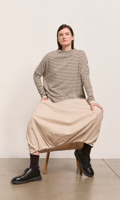 Plümo  Person in Oana Mock Neck striped sweater and beige balloon skirt sits on a wooden stool against a plain backdrop.