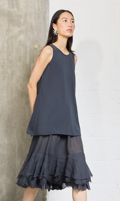 Plümo- Woman in a relaxed fit, sleeveless Edel Top and ruffled skirt stands against a concrete and white wall, flower earrings on.