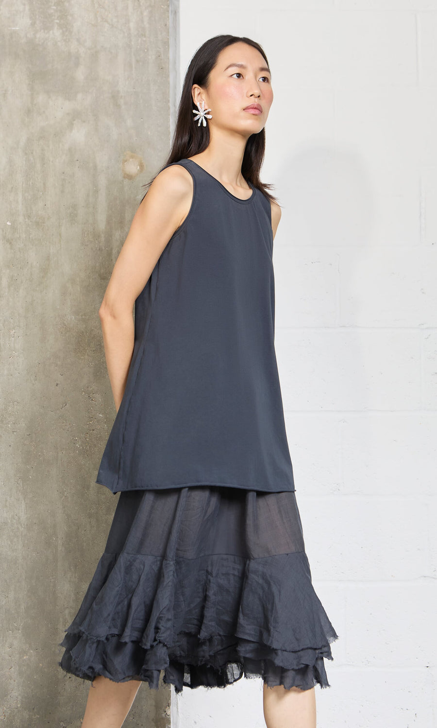 Plümo- Woman in a relaxed fit, sleeveless Edel Top and ruffled skirt stands against a concrete and white wall, flower earrings on.