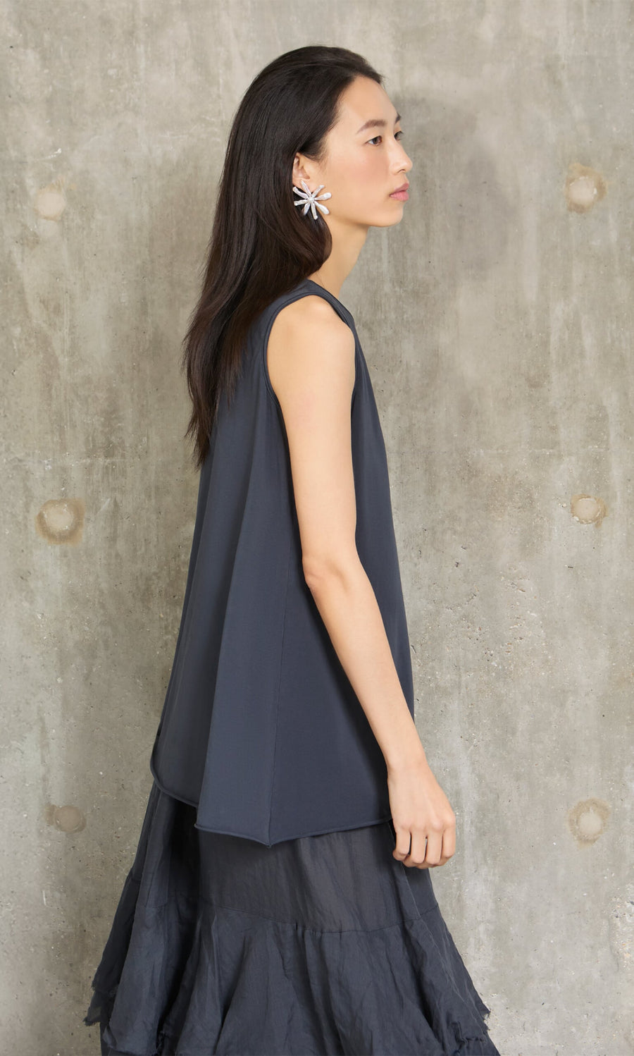 Plümo- Woman in the Edel Top, a relaxed fit organic cotton tank, with a ruffled skirt and floral earring by a concrete wall.