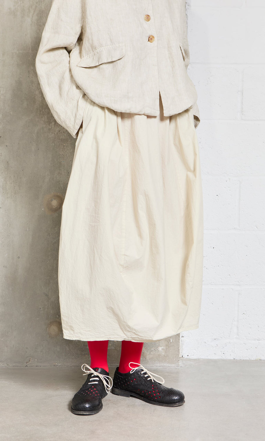 Plümo- Wearing the Edina Skirt, a beige jacket, red socks, and black lace-up shoes, they stand by a concrete wall.