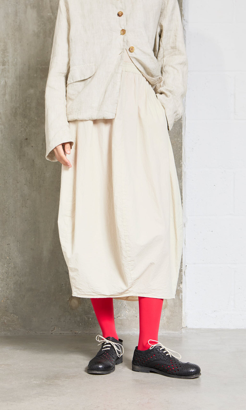 Plümo- Person wearing the Edina Skirt with red tights and black lace-up shoes, standing against a concrete wall.