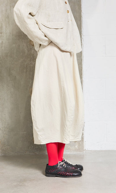 Plümo- Person in the cream Edina Skirt, bright red tights, and black shoes stands against a concrete and white wall.