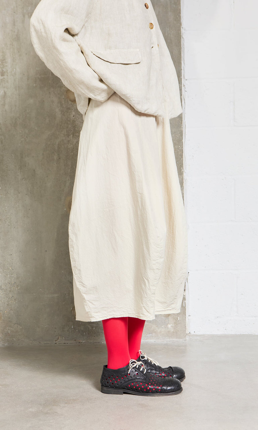 Plümo- Person in the cream Edina Skirt, bright red tights, and black shoes stands against a concrete and white wall.