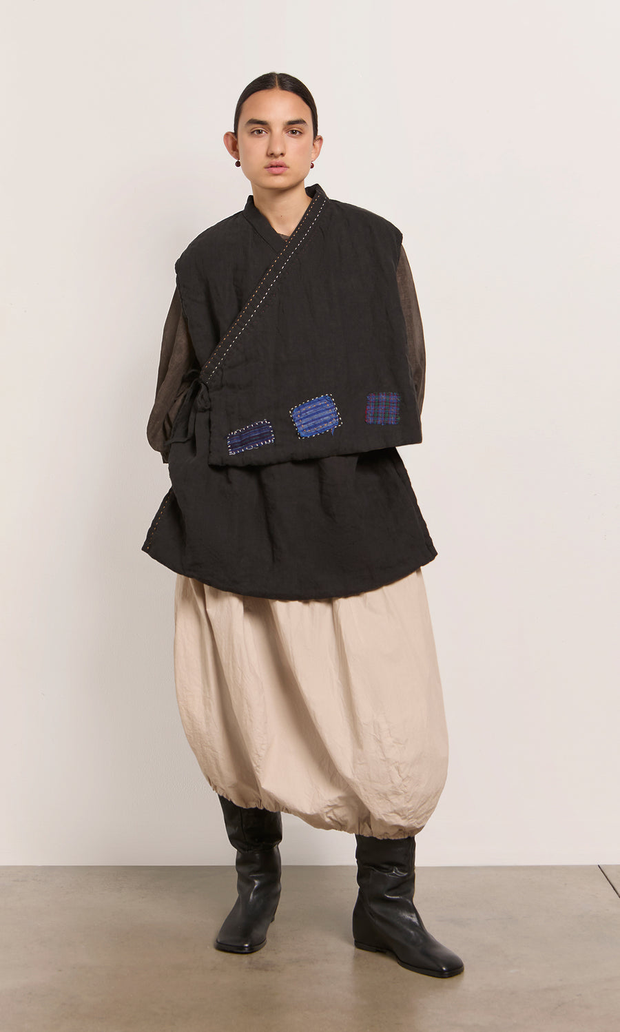 Plümo  Model wears Engi Waistcoat, loose beige pants, and black boots, standing by a plain wall.