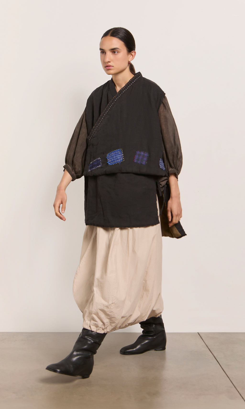 Plümo  Person in loose beige pants, black boots, sheer-sleeved top, and the Engi Waistcoat with handstitched patches walks indoors.