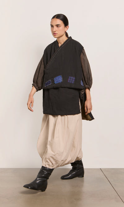 Plümo  Person in loose beige pants, black boots, sheer-sleeved top, and the Engi Waistcoat with handstitched patches walks indoors.