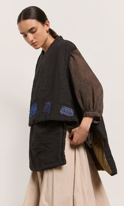 Plümo  Woman in the Engi Waistcoat—black linen with blue handstitched patches, brown sheer sleeves, cream skirt, looking down.