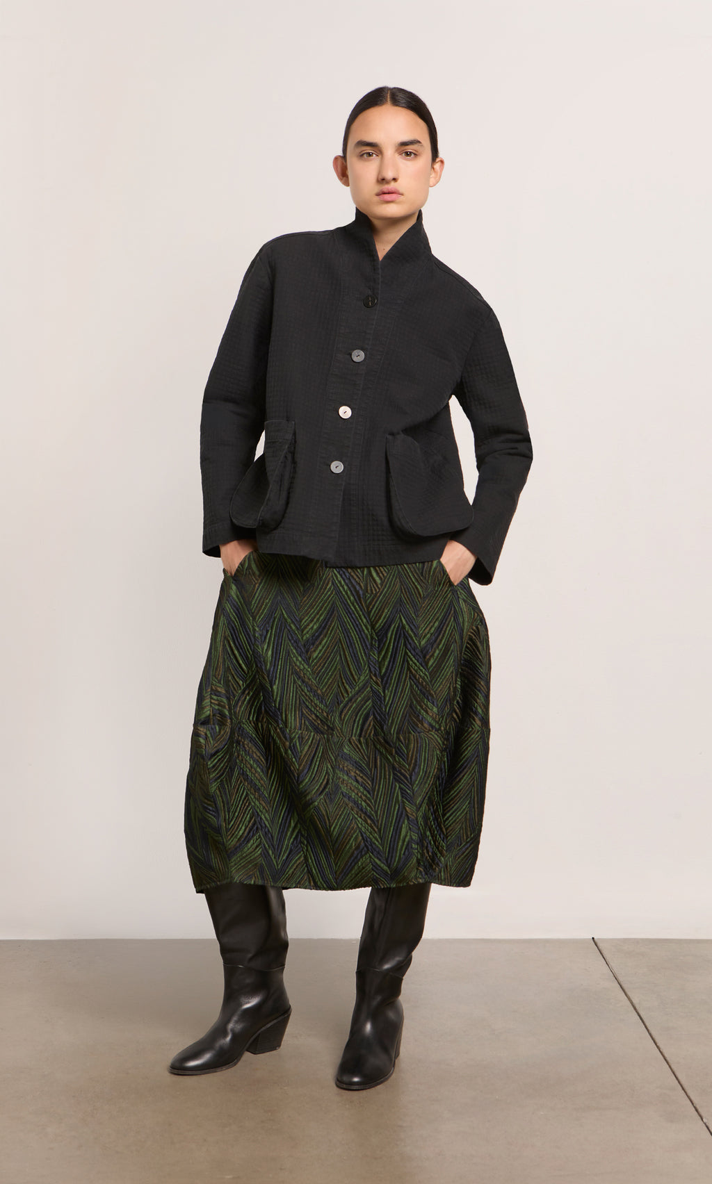 Plümo  Woman in a black jacket, Yona Skirt, and tall black boots stands against a plain background.