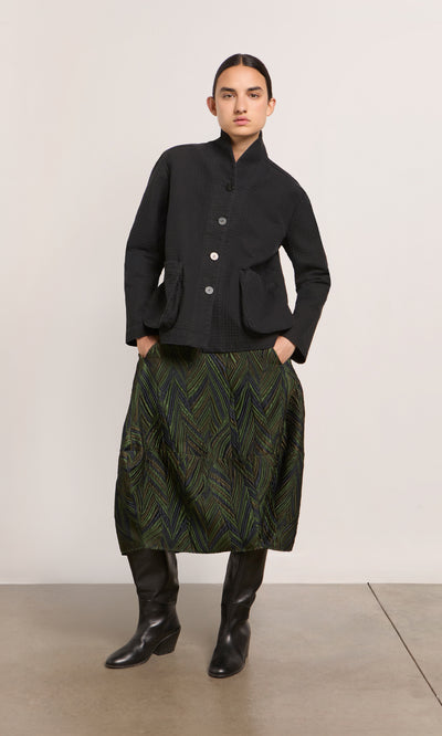 Plümo  Woman in a black jacket, Yona Skirt, and tall black boots stands against a plain background.
