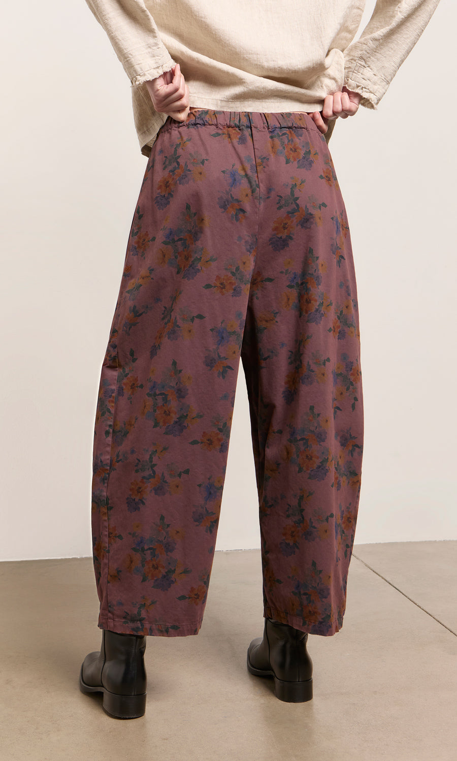 Plümo  Model wears Espel Pants in purple floral, a cream top, and black boots on a light floor, facing away.