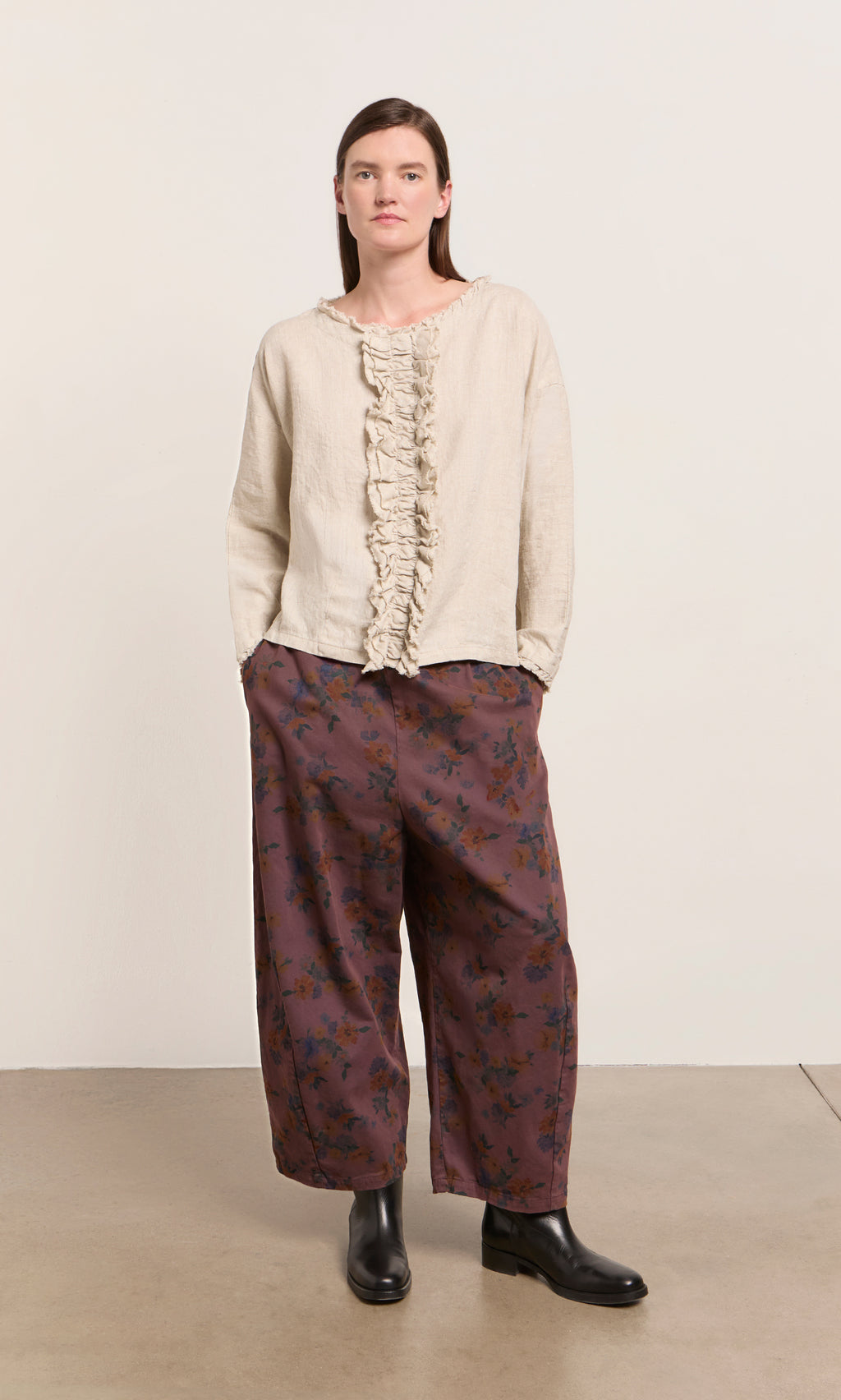 Plümo  Someone in a beige ruffled top, Espel Pants with floral print, and black boots stands by a plain light background.