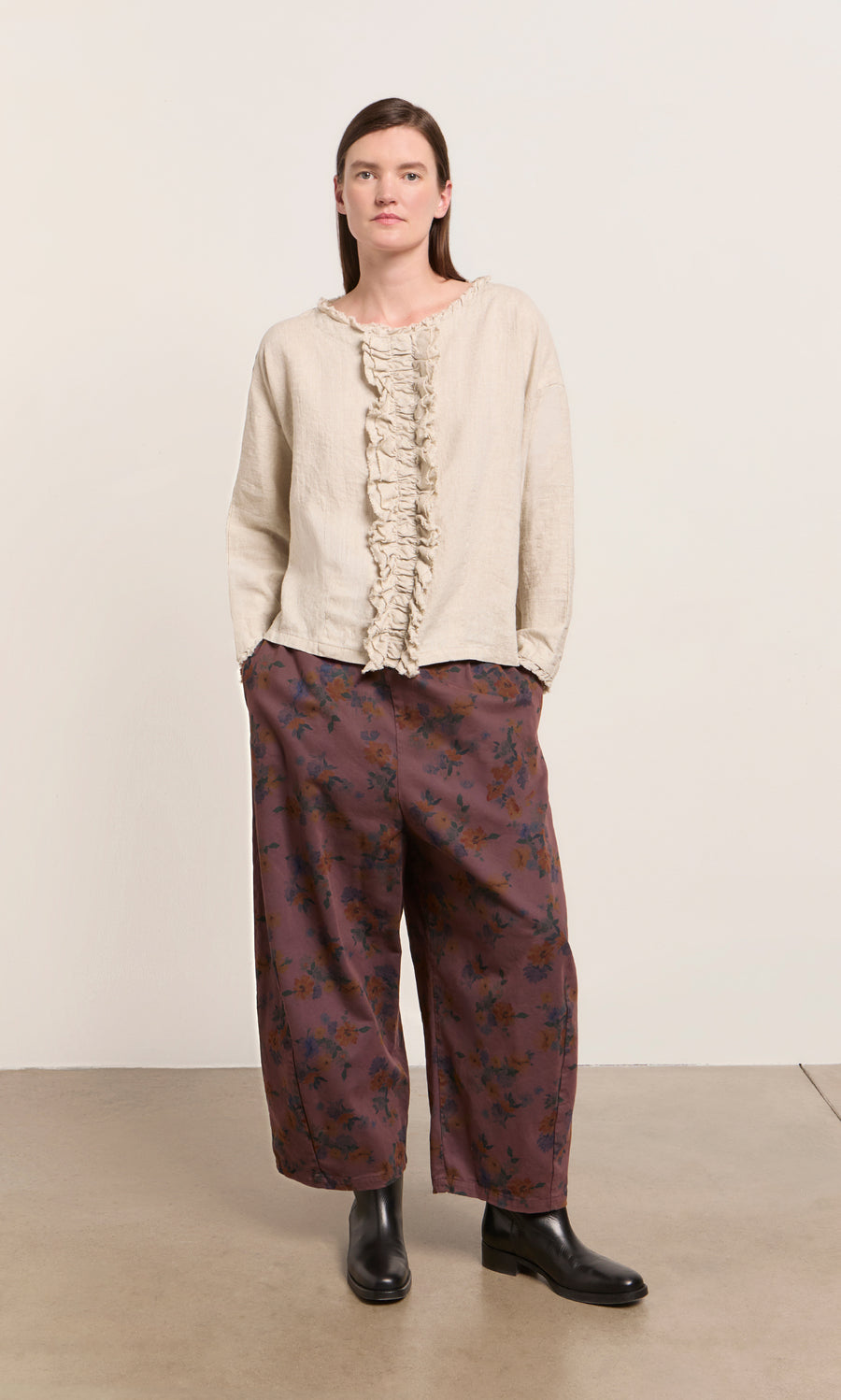 Plümo  Someone in a beige ruffled top, Espel Pants with floral print, and black boots stands by a plain light background.
