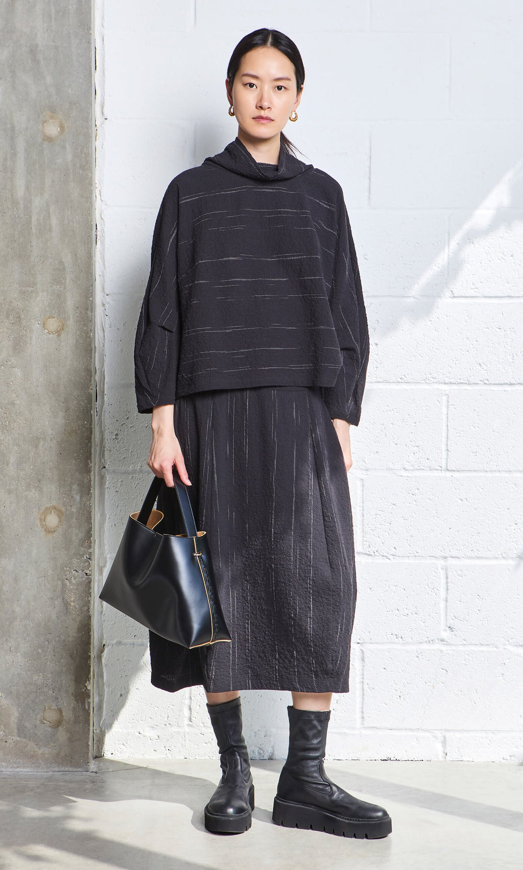 Plümo- Woman in a cowl neck blouse, black skirt and boots, holding a bag, standing against a white wall.
