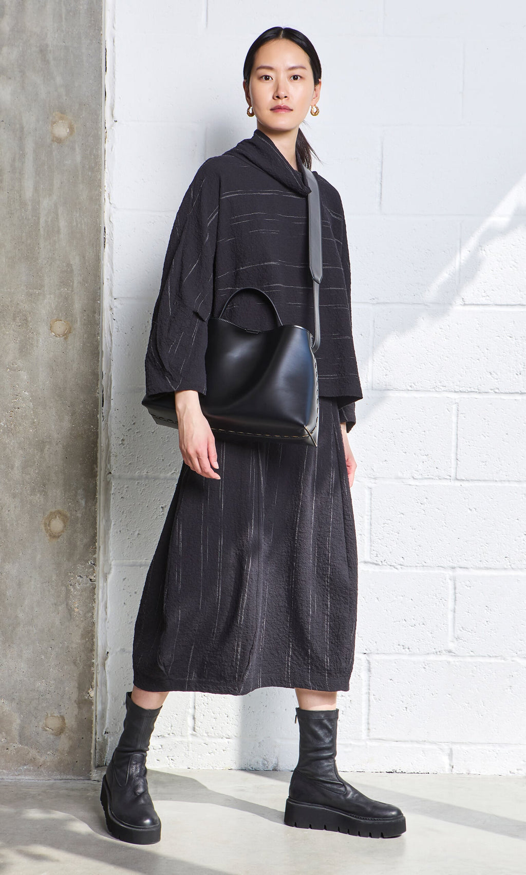 Plümo- Woman in the Speckle Skirt, ankle boots, and black handbag stands against a white brick wall.