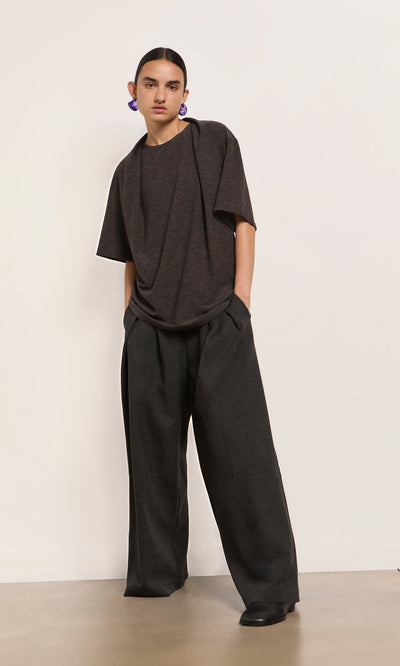 Plümo  Person in Feld Tee, loose dark pants, and purple earrings stands against a plain light wall.