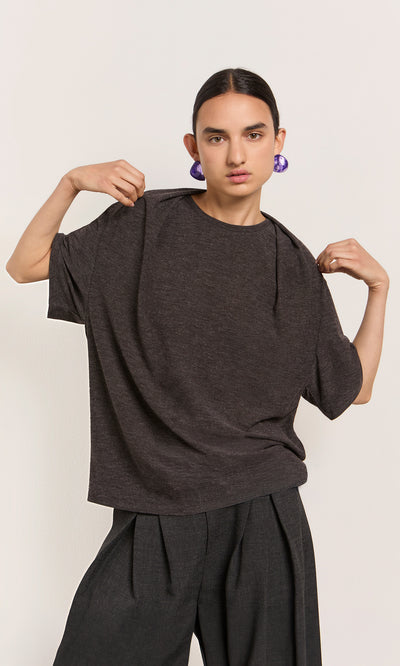 Plümo  A model wears the Feld Tee, a marl grey jersey boxy top, with hair pulled back, purple earrings, and pleated pants.
