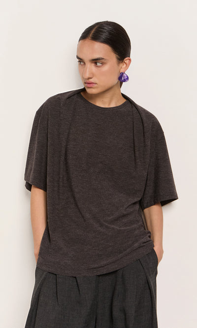 Plümo  A person with dark hair in a low bun wears a marl grey Feld Tee, wide pants, and large purple earrings.