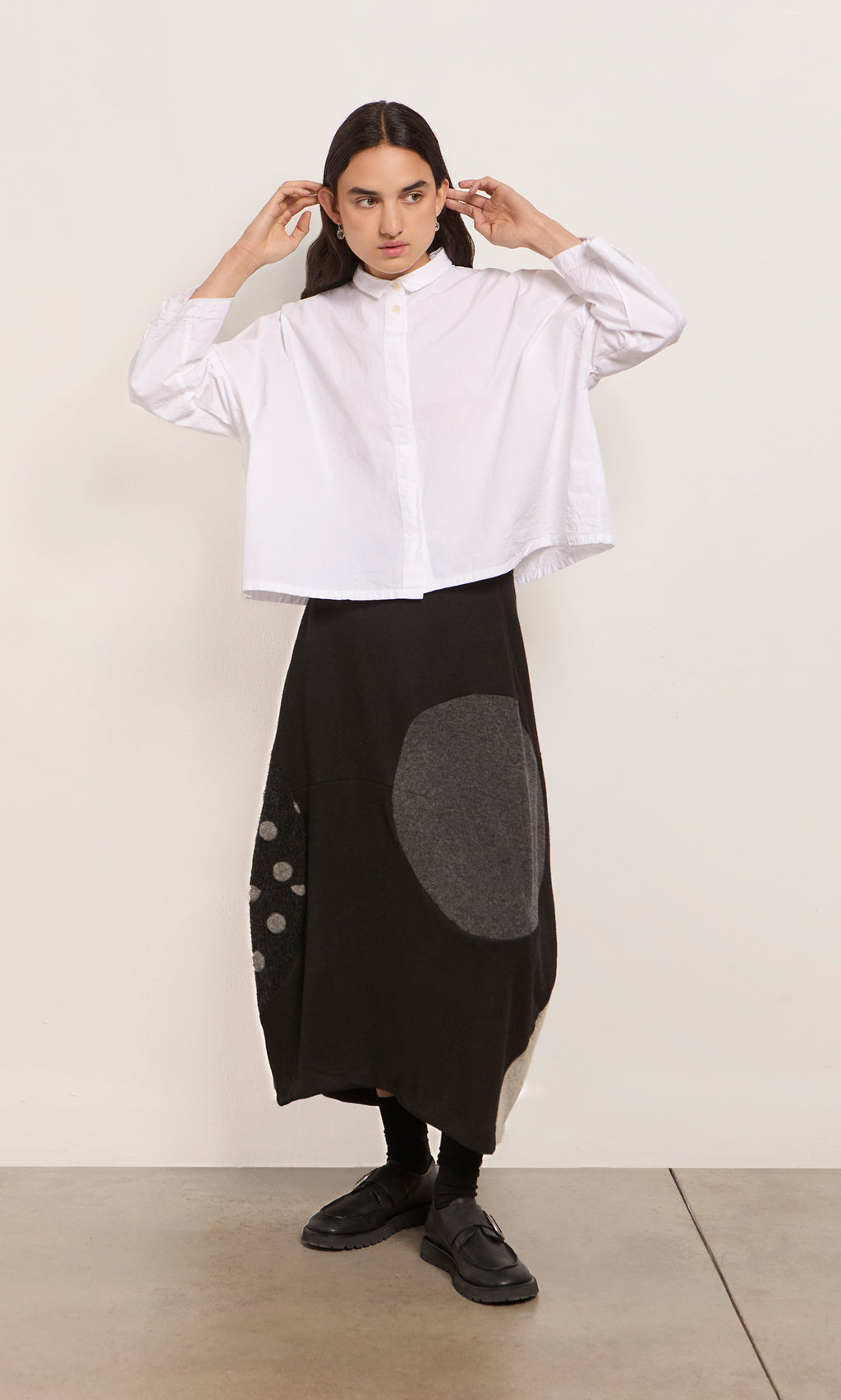 Plümo  Person in a white button-up shirt and black patchwork skirt stands against a plain light wall.