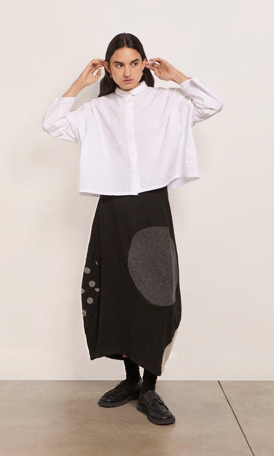 Plümo  Person in a white button-up shirt and black patchwork skirt stands against a plain light wall.