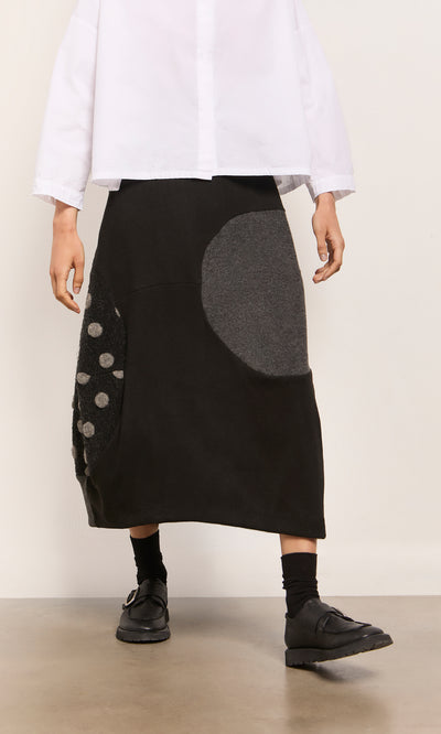 Plümo  Person in white shirt, Felted Bubble Skirt with gray circles, black shoes and socks, standing indoors.