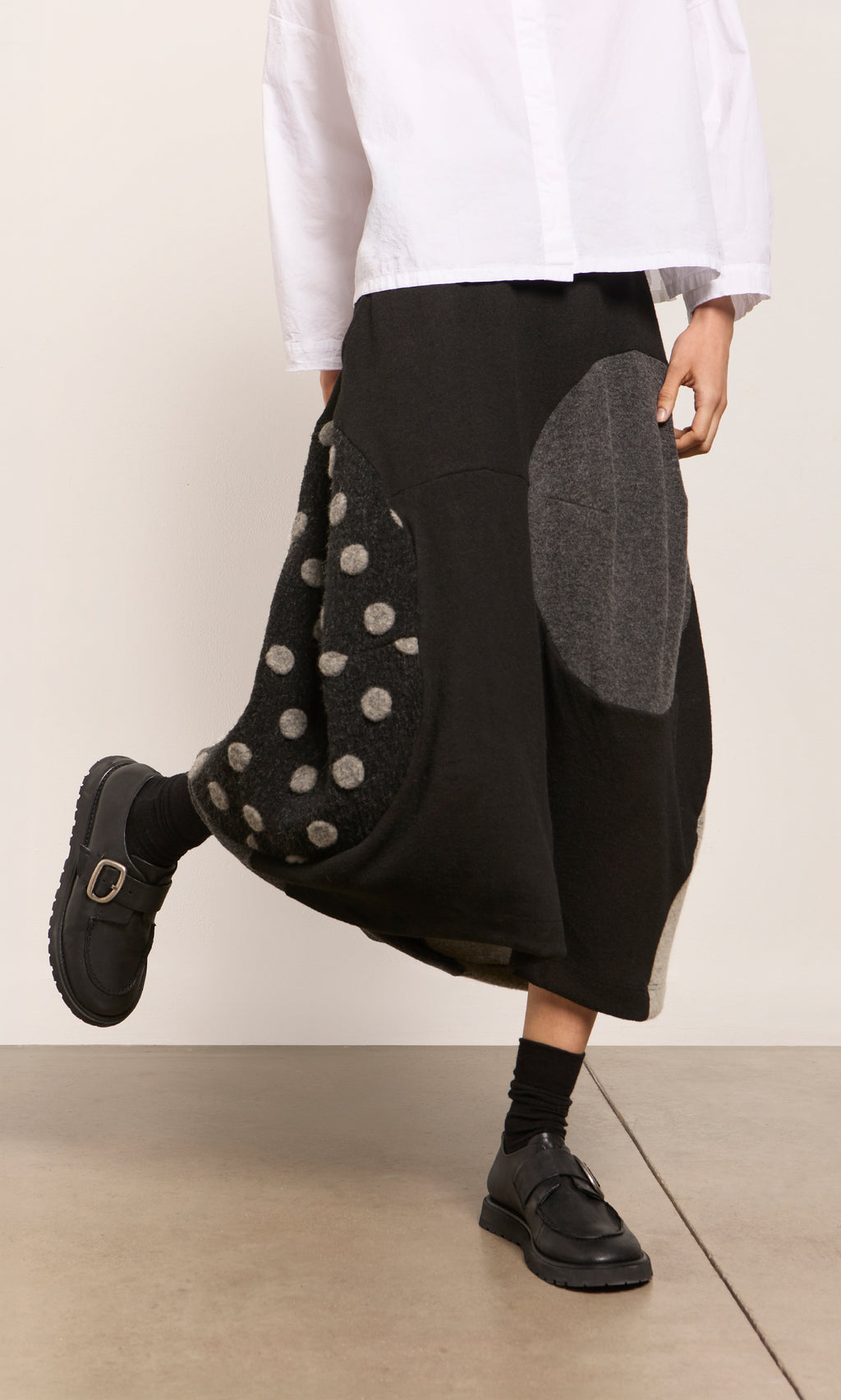 Plümo  Person in a white shirt, black Felted Bubble Skirt with circles, black socks and shoes on a light floor.