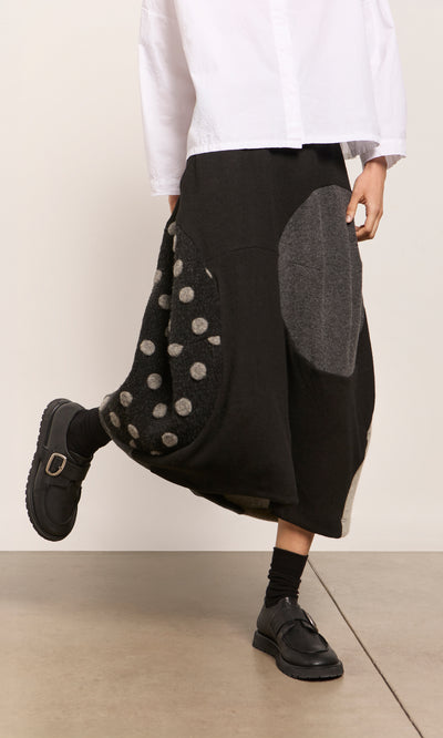 Plümo  Person in a white shirt, black Felted Bubble Skirt with circles, black socks and shoes on a light floor.