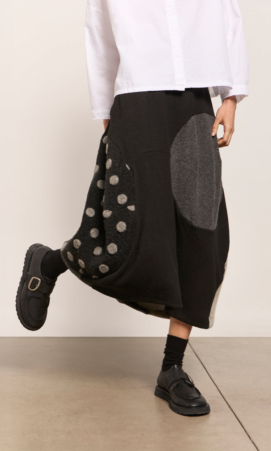 Plümo  Person in a white shirt, black Felted Bubble Skirt with circles, black socks and shoes on a light floor.