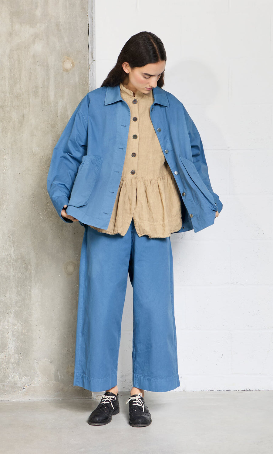 Plümo- A woman stands indoors in Cromer Pants - French Blue with a blue jacket over a beige top, looking down at her outfit.