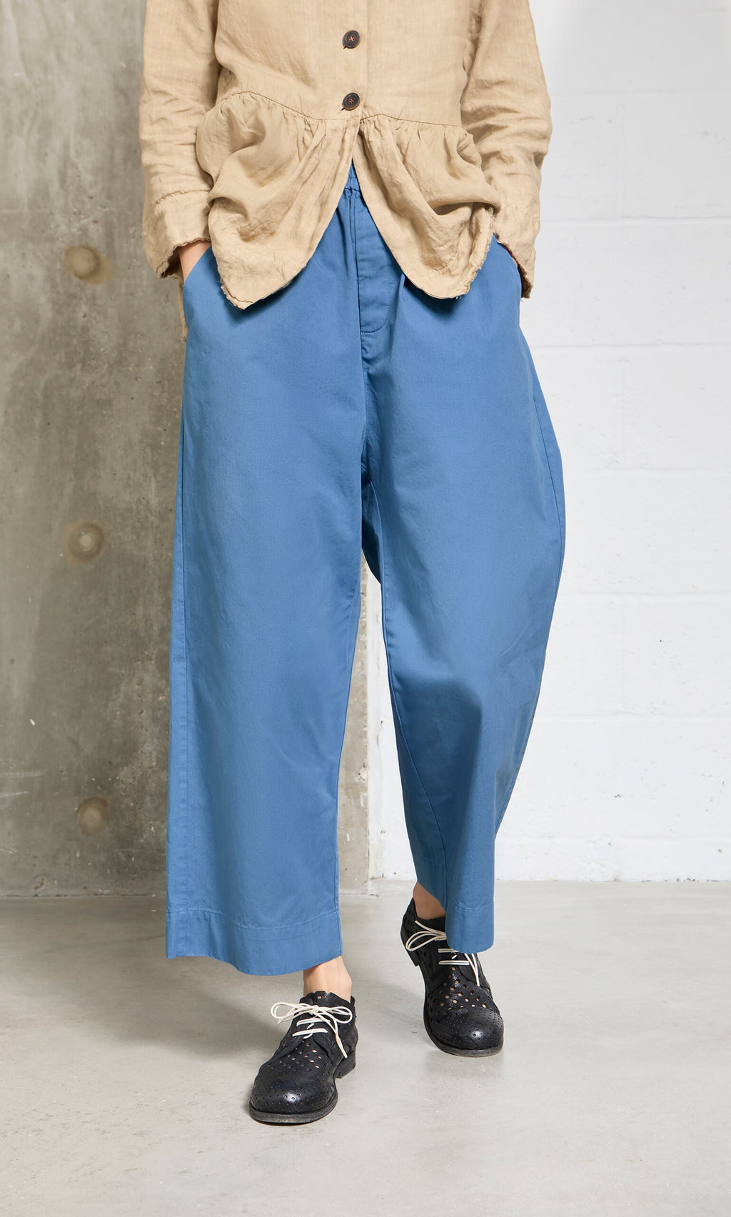 Plümo- Person in a beige top and Cromer Pants - French Blue, indoors on concrete floor, wearing black shoes with white laces.